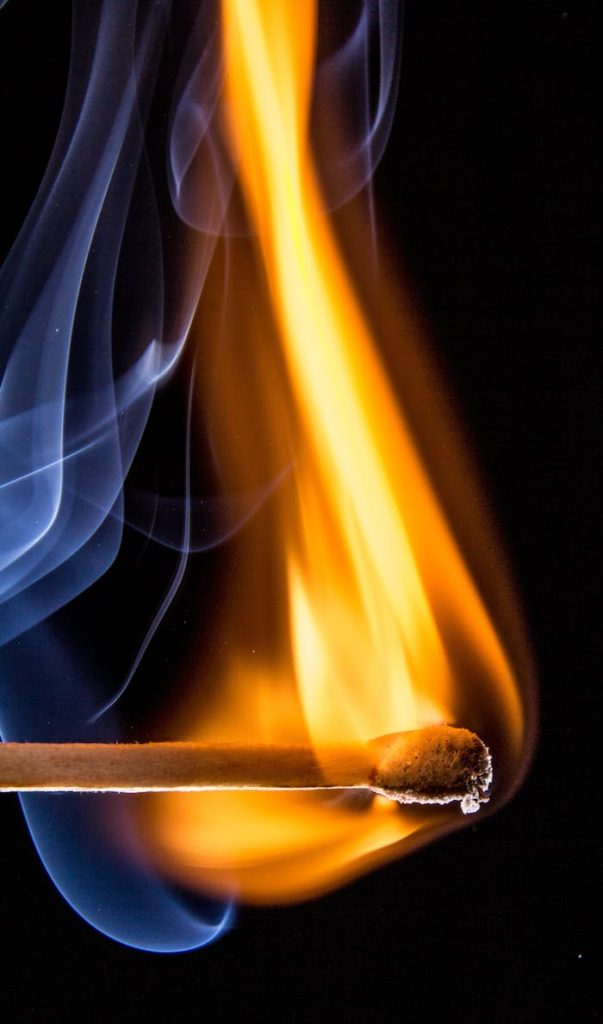 Dramatic close-up of a matchstick igniting with vivid orange flames and wisps of smoke.